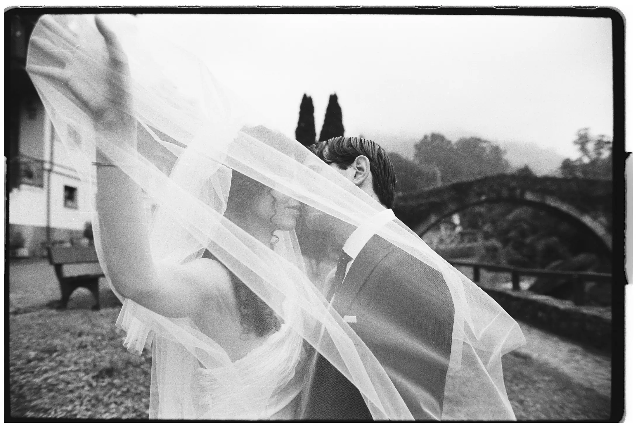 Foto en blanco y negro de una pareja vestida de boda. Ella sujeta el velo delante de ellos que están dándose un beso. De fondo se ve un puente antiguo y un río.